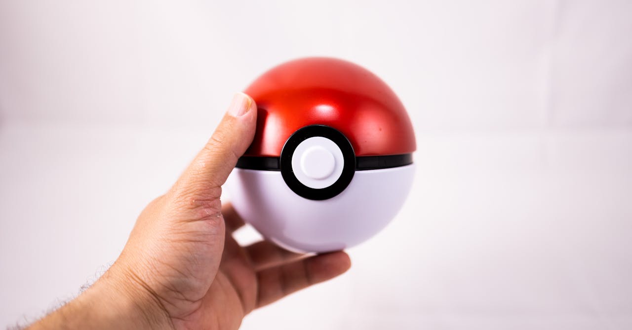 Close-up of a human hand holding a red Pokeball toy against a white background.